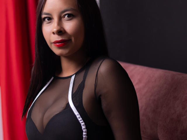 Clair Bennet, con cabello oscuro, lápiz labial rojo, una blusa negra transparente que deja ver sus senos, sentada frente a un fondo rojo y negro, sonriendo suavemente..