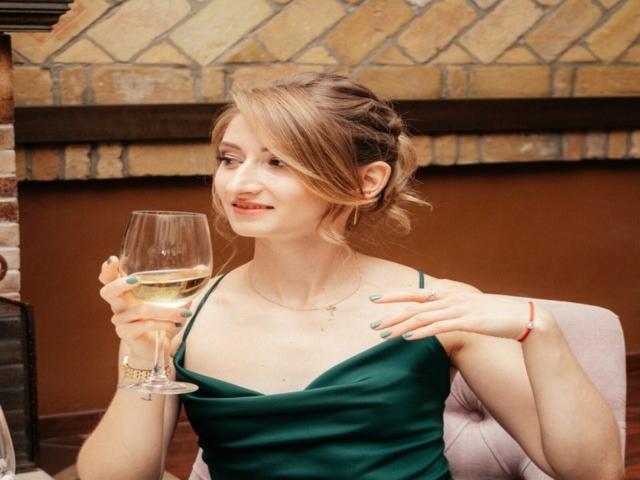 GwynBartell with light brown hair in a bun, green dress, holding a glass of white wine, smiling, hand on chest, pink chair, red bracelet, green nails.