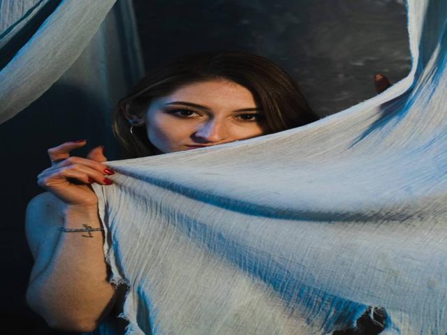 GwynBartell with dark brown hair, red nails, and a tattoo on her right wrist, peeks from behind a torn, light blue fabric, showing her face and upper chest.