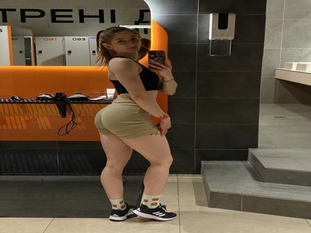 GwynBartell mit einem hübschen Hintern, trägt ein schwarzes Crop-Top, beige gerippte Shorts, weiße Socken mit goldenen Mustern und schwarze Sneakers und macht ein Selfie vor einem Spiegel im Badezimmer..