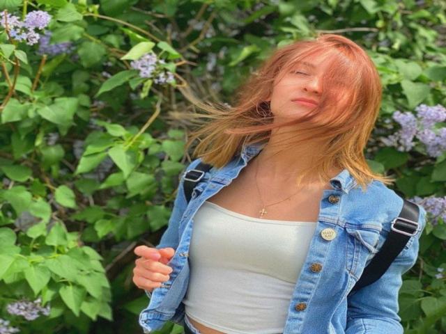 GwynBartell hat hellbraunes Haar, eine blaue Jeansjacke, ein weißes Top und eine Kreuzkette. Sie steht mit einem schwarzen Träger über der Schulter, umgeben von grünen Blättern und lila Blumen..