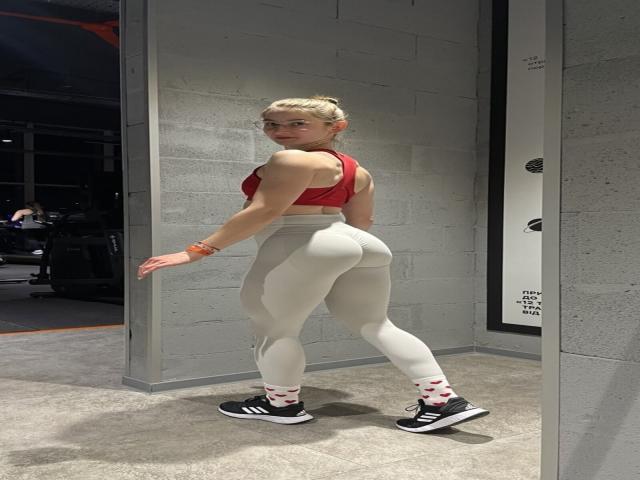 GwynBartell, avec des cheveux blonds attachés en chignon, un soutien-gorge de sport rouge, un legging blanc taille haute, des baskets noires avec des chaussettes blanches et rouges, debout, avec une fesse proéminente, regardant en arrière..