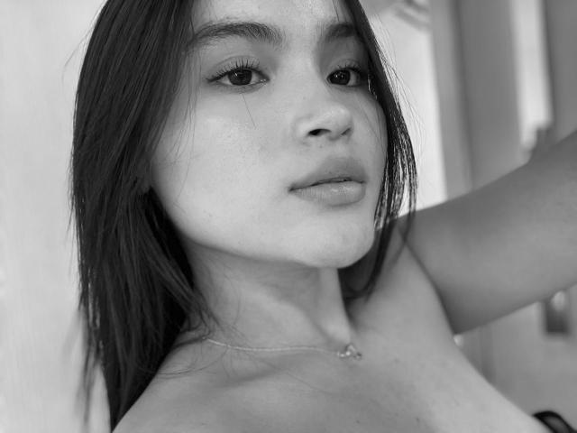 Black and white photo of MiiaPalmer with medium-length dark hair, freckled face, subtle makeup, and a necklace, showing her bare shoulders and a relaxed, confident expression.