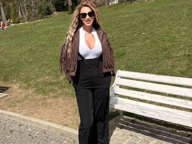 NicolSNaya with wavy blonde hair, wearing black pants, white halter top, brown fringed jacket, and black sunglasses, standing by a white wooden bench on a sunny day.