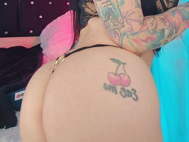 Kkole's tattooed butt with a cherry and "umm 03" on her right cheek, wearing a black thong, showing colorful arm tattoos, and pink hair.