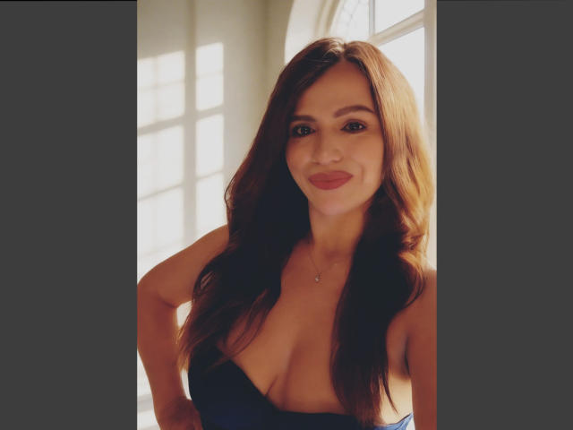 CameronViviane with long brown hair, smiling, wearing a low-cut black dress, showing her tits, standing with hands on hips, sunlight through window.