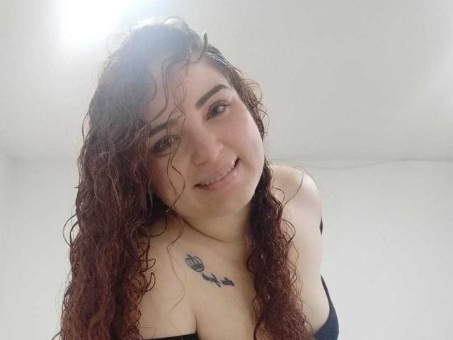 AmandaWt with curly brown hair, smiling down, showing her tits, has a tattoo of a flower and leaves on her chest, in a white room.