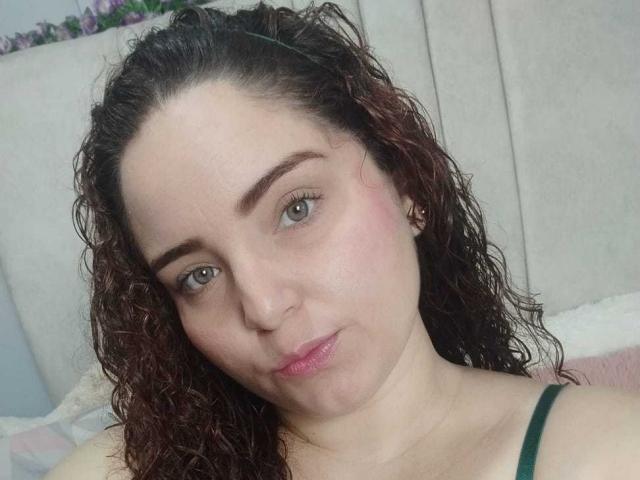 AmandaWt with curly brown hair, green eyes, pink lips, and light skin, wearing a green strap, looking at the camera with a subtle smile.
