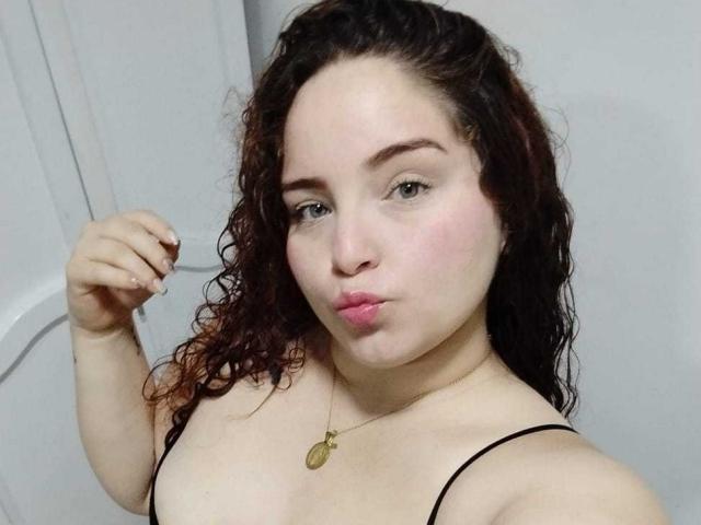 AmandaWt mit lockigem, braunem Haar, heller Haut und rosafarbenen Lippen, die leicht zusammengepresst sind, trägt ein schwarzes Top mit Trägern, eine goldene Halskette und hat die Faust geballt. Ihre Brüste sind leicht sichtbar..