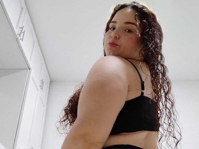 AmandaWt, con cabello castaño largo y rizado, mirando por encima del hombro, vistiendo un sostén negro, mostrando su espalda y hombro desnudos, senos pequeños y una ligera expresión de puchero en su rostro..
