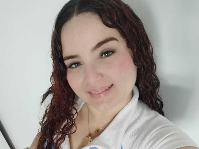 AmandaWt has curly brown hair, green eyes, and a slight smile. She's wearing a white shirt, showing her cute tits, and a gold necklace with a heart pendant.