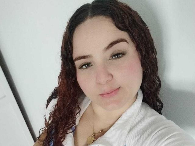 AmandaWt has curly brown hair, light skin, and green eyes. She's wearing a white shirt, smiling slightly, with a gold necklace. Her boobs are partially visible.