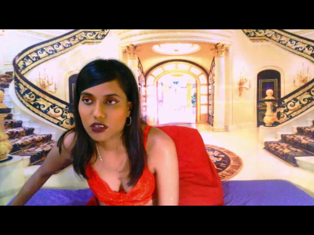 IndianSkies with dark hair and red dress, leaning on purple bed, showing tits, with purple lipstick and hoop earrings, in a luxurious room with ornate stairs.