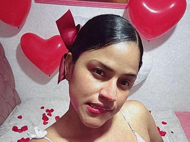 IvannaDuran with dark hair in a red bow, pink lips, and a slightly open mouth, wearing a white top, surrounded by red heart balloons and rose petals.