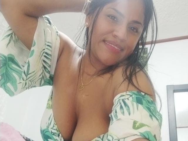 IvannaDuran with medium brown skin, long black hair, smiling, wearing a green leaf-patterned off-shoulder top, revealing her large tits, gold necklace, and pink lipstick.