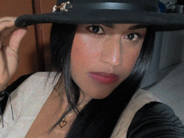 IvannaDuran with black hair, wearing a black hat and pink lipstick, holding the hat with her right hand, wearing a beige top with a black jacket, looking at camera.