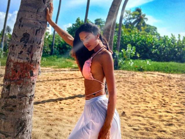 VicktoriaSunshine stands by a palm tree, wearing a pink bikini top and white skirt, with tan skin, long dark hair, and a slim, attractive ass. Sunlight filters through.