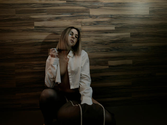 IceMaya with glasses, blonde bob, white shirt open showing her tits, black skirt, sitting against wood panel wall, holding bottle, dark moody lighting, sexy, confident.