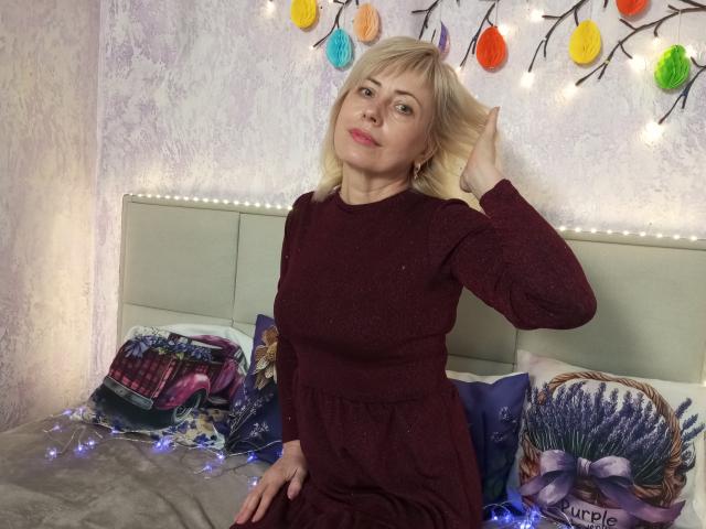 LaurenWishy, blonde, wearing a maroon dress, sits on a bed with colorful pillows and string lights, hand in hair, smiling, looking at camera, medium tits, cute.