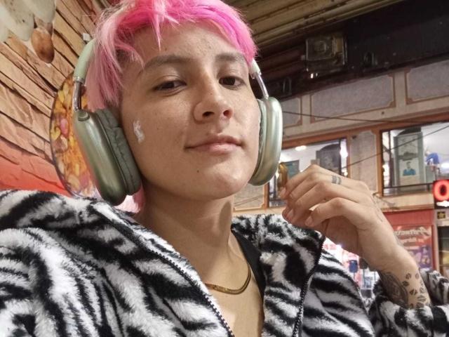 DannyRouds with pink hair, wearing zebra-patterned hoodie, silver headphones, slight smile, tattoo on right wrist, small white mark on cheek, gold necklace, indoors.