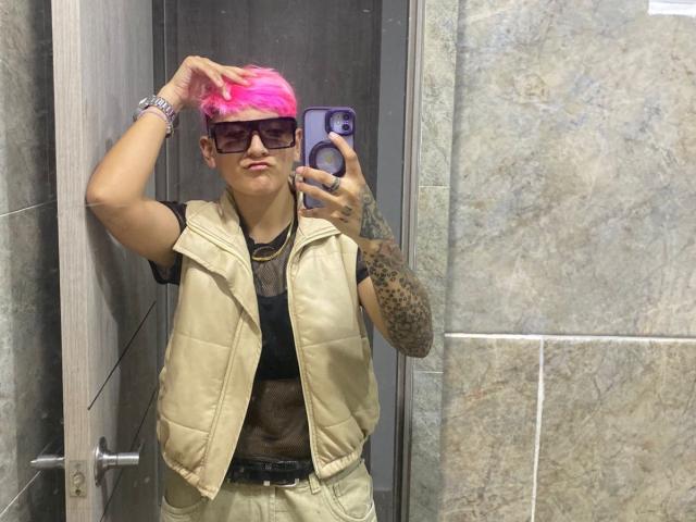 DannyRouds with pink hair, tattoos, wearing a beige vest, black shirt, and sunglasses, taking a mirror selfie in a bathroom. His hand is in his hair.