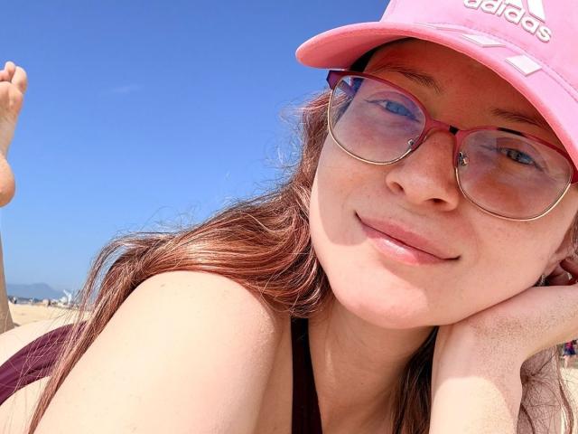 AmbeerWood with pink Adidas cap, pink sunglasses, and brown hair, smiling at the camera, lying on a sunny beach, showing freckles and a relaxed, cute face.