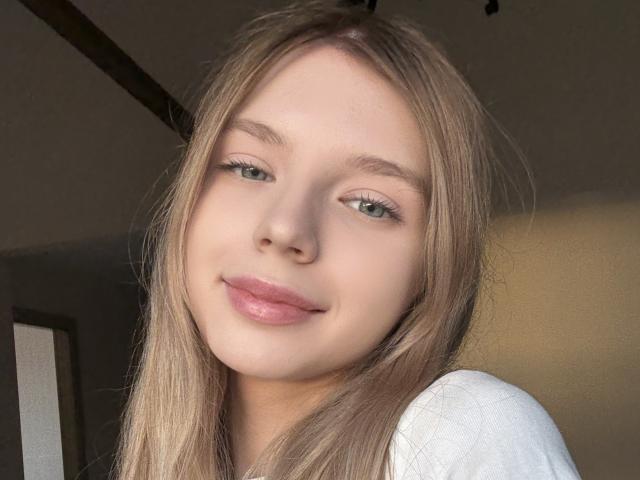 BernnyChloe has light blonde hair, green eyes, and pink lips with a slight smile. She's wearing a white shirt, and the photo is a cute selfie.