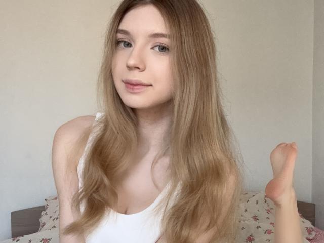 BernnyChloe with long, straight blonde hair, wearing a white tank top, showing a small, pale breast, and a raised foot in a casual, light-filled setting.