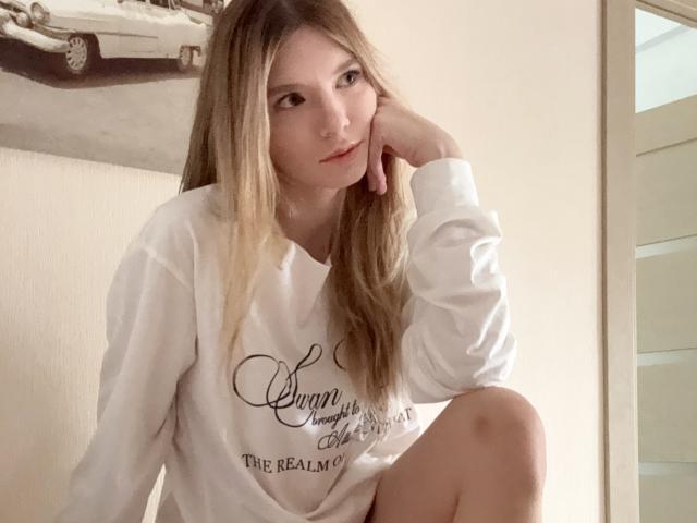 BernnyChloe with long blonde hair, white long-sleeve shirt, and subtle makeup, resting her head on her hand, sitting with one knee up, looking at the camera.