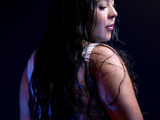 BarbaraHoneyy with wet black hair, eyes closed, wearing a white tank top, showing her ass, against a dark background, illuminated by blue and purple light.