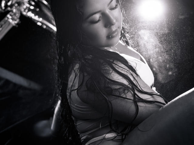 Black and white photo of BarbaraHoneyy with wet, long hair, eyes closed, wearing a wet white shirt, light shining from the right, showing her tits and slightly visible ass.
