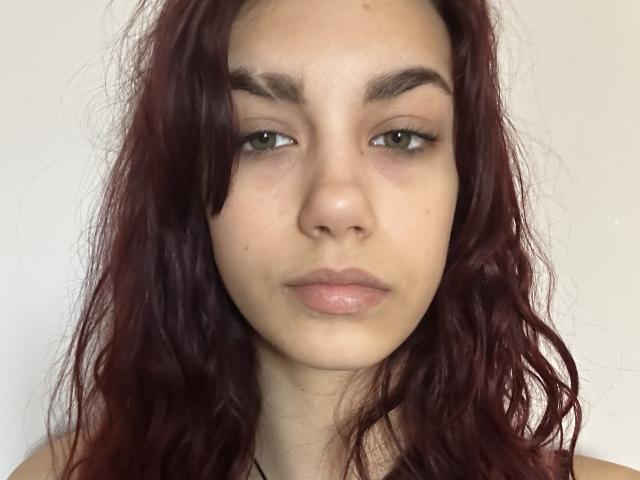 AuroraElly has wavy, dark red hair, green eyes, and light skin with a neutral expression. She's showing her pouty, glossy pink lips and slightly thick eyebrows.