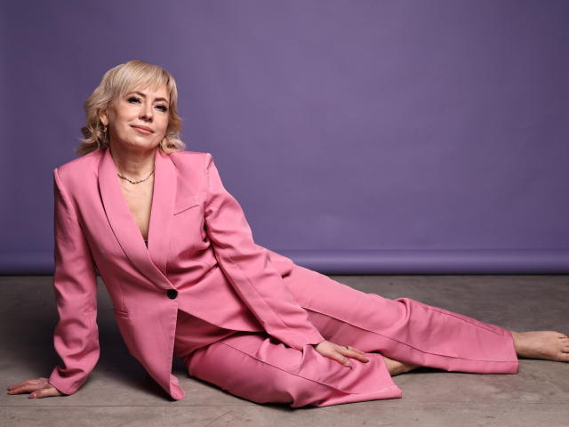 LaurenWishy, blonde, in a pink suit, sitting on the floor, legs stretched out, hand on knee, smiling, purple background, barefoot, pink jacket, pink pants, subtle makeup.