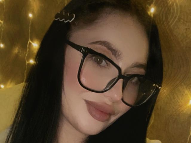 KristyAnita, com óculos pretos, cabelo escuro e um sorriso discreto, tem um rosto adorável, lábios carnudos e um leve rubor, usando uma presilha de cabelo no lado esquerdo..