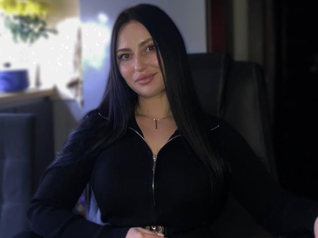 KristyAnita tiene el pelo largo y negro, pechos de tamaño medio, y lleva una camiseta negra con cremallera y un collar con una cruz, está sentada en una silla oscura y sonríe suavemente..