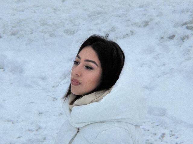 DeannDelcamp with dark hair, white puffy coat, and beige scarf, looking over her shoulder, standing in snow, showing her pouty, pink-lipped face.