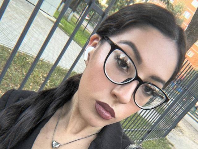 DeannDelcamp with dark brown hair, glasses, and purple lipstick, wearing a black jacket and necklace, has white earbuds in. Her face is slightly turned, looking at the camera.