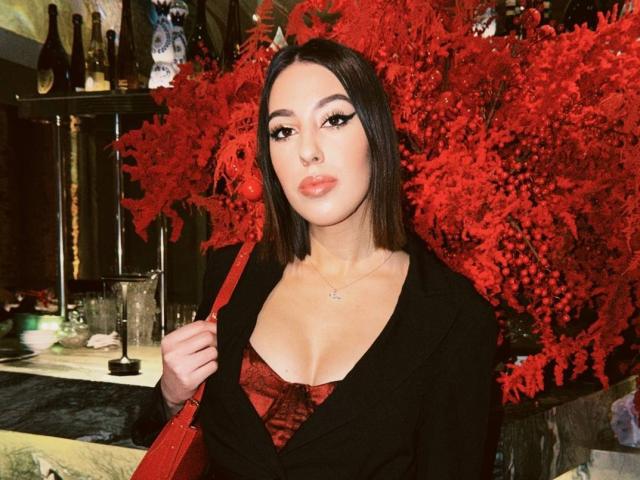 DeannDelcamp with dark straight hair, wearing a black blazer, red lace-up bustier, showing her tits, red bag, and dramatic eyeliner, standing in front of red foliage.