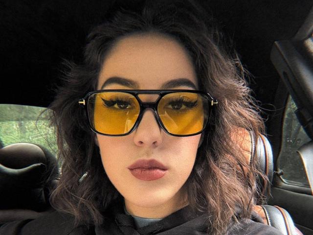 DeannDelcamp with curly dark hair, wearing black tortoiseshell sunglasses with yellow lenses, looking straight at the camera, sitting in a car with a black seat.