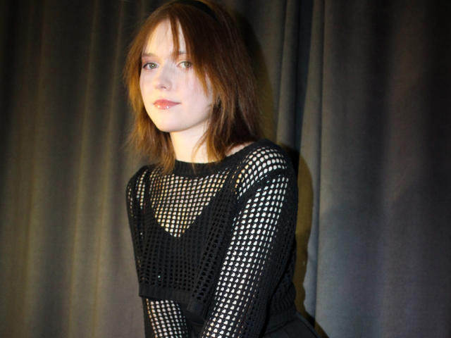 YukiKendal with shoulder-length brown hair, wearing a black mesh long-sleeve top, exposing her small tits, standing against a dark curtain, looking slightly to the side.