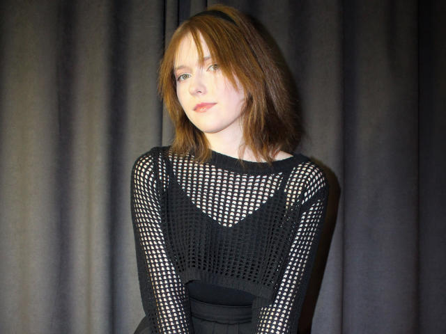 YukiKendal with pale skin, brown hair, and a black mesh top showing her small tits. She's looking to the side with a slight smile, standing against dark curtains.