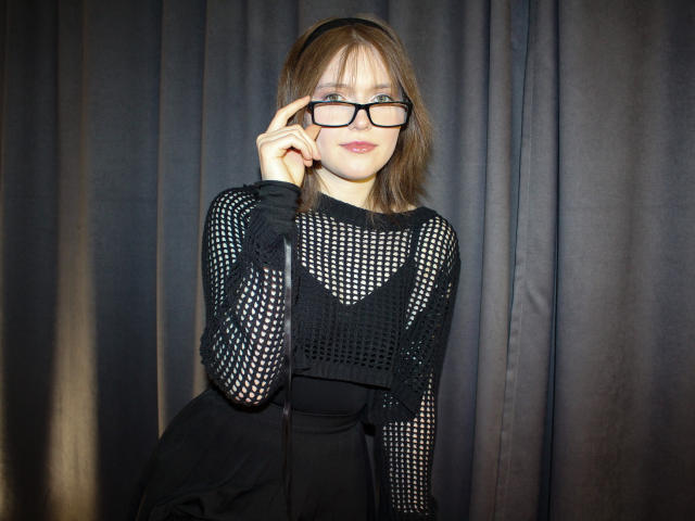 YukiKendal with glasses, black mesh top, and black skirt, touching her glasses, smiling, brown hair, fair skin, standing against dark curtains, showing her tits slightly through mesh.