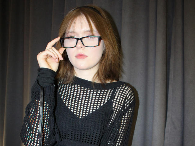 YukiKendal with brown hair, wearing black mesh top, black-framed glasses, and black headband, touching glasses with right hand, looking slightly annoyed, pale skin, small tits.