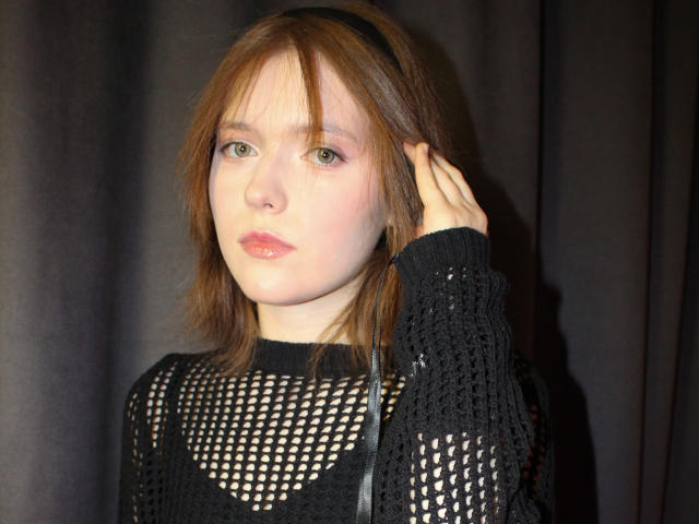 YukiKendal has pale skin, green eyes, and messy brown hair with a black headband. She's wearing a black, mesh, knit top, looking serious with a hand in her hair.