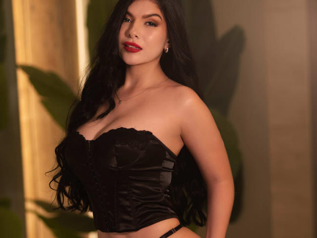 ManuelaOcampoz with long black hair, wearing a black lace-up corset, showcasing her large, round, firm boobs, and red lipstick, standing in a warm, softly-lit setting.
