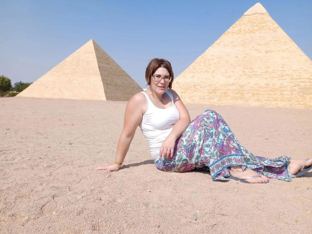 ZimtStern-hot mit braunen Haaren, Brille, weißem Tanktop und Blumenhose sitzt auf Wüstensand, lehnt sich zurück und zeigt ihren Hintern, im Hintergrund sind zwei Pyramiden zu sehen, der Himmel ist klar..