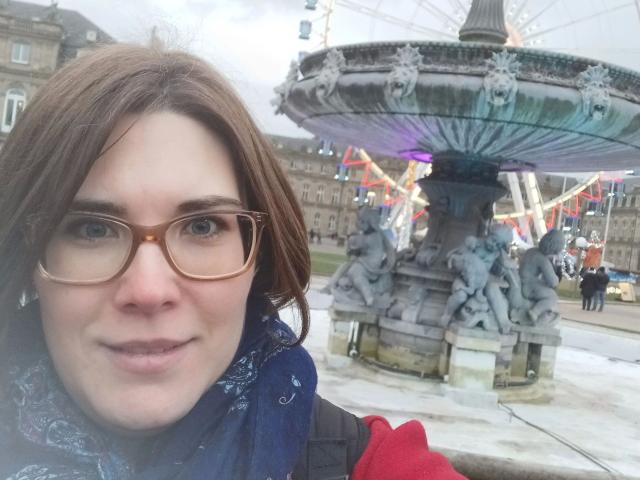ZimtStern-hot has brown hair, glasses, and a blue scarf, standing in front of a fountain with statues. Her red jacket is visible. The fountain is in the background.