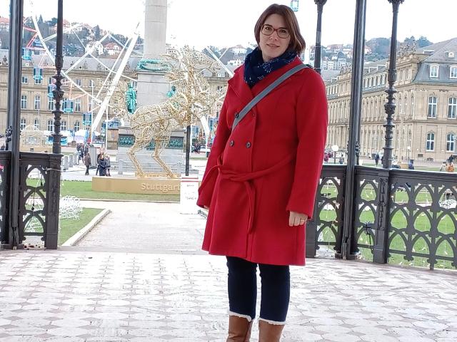 ZimtStern, vestito con un cappotto rosso, pantaloni neri e stivali marroni, in piedi su un ponte di pietra, con una sciarpa blu e occhiali, che guarda leggermente di lato..