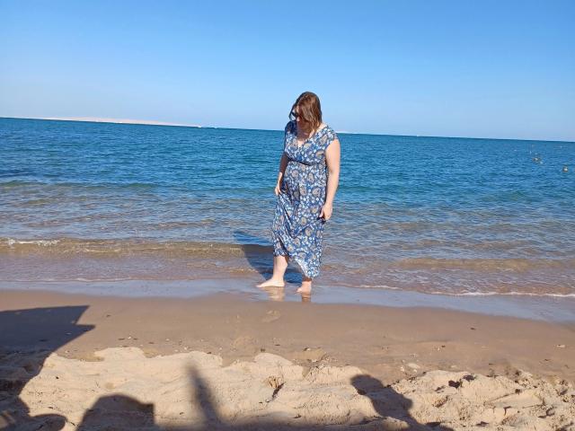 ZimtStern trägt ein blaues Kleid mit Blumenmuster, ist barfuß und geht am Strand entlang, wo sanfte Wellen brechen. Sie blickt nach unten, und das Sonnenlicht wirft Schatten..
