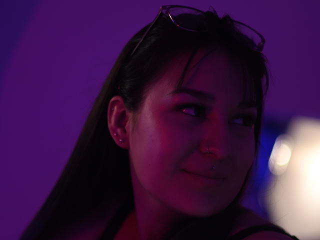 LunaColliins, avec ses cheveux foncés, son piercing au nez et ses petits boucles d'oreilles, porte des lunettes de soleil sur la tête, dans une lumière violette-bleue tamisée, et sourit légèrement. Ses seins ne sont pas visibles..